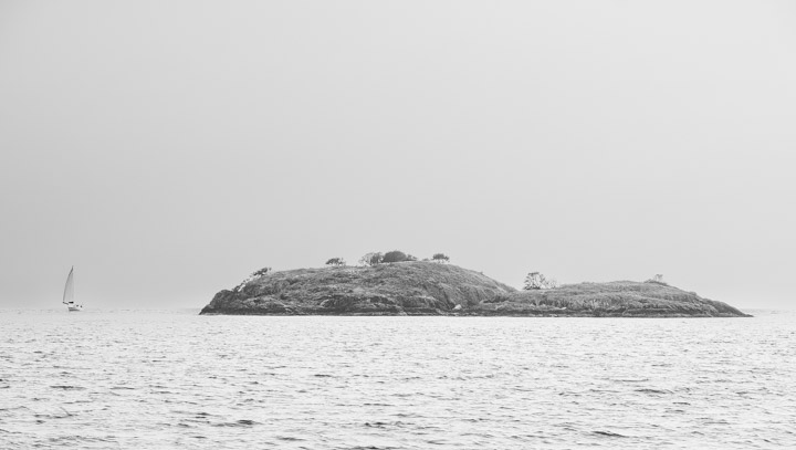 Solitary rock in Átl’ḵa7tsem, sailboat off to the side.